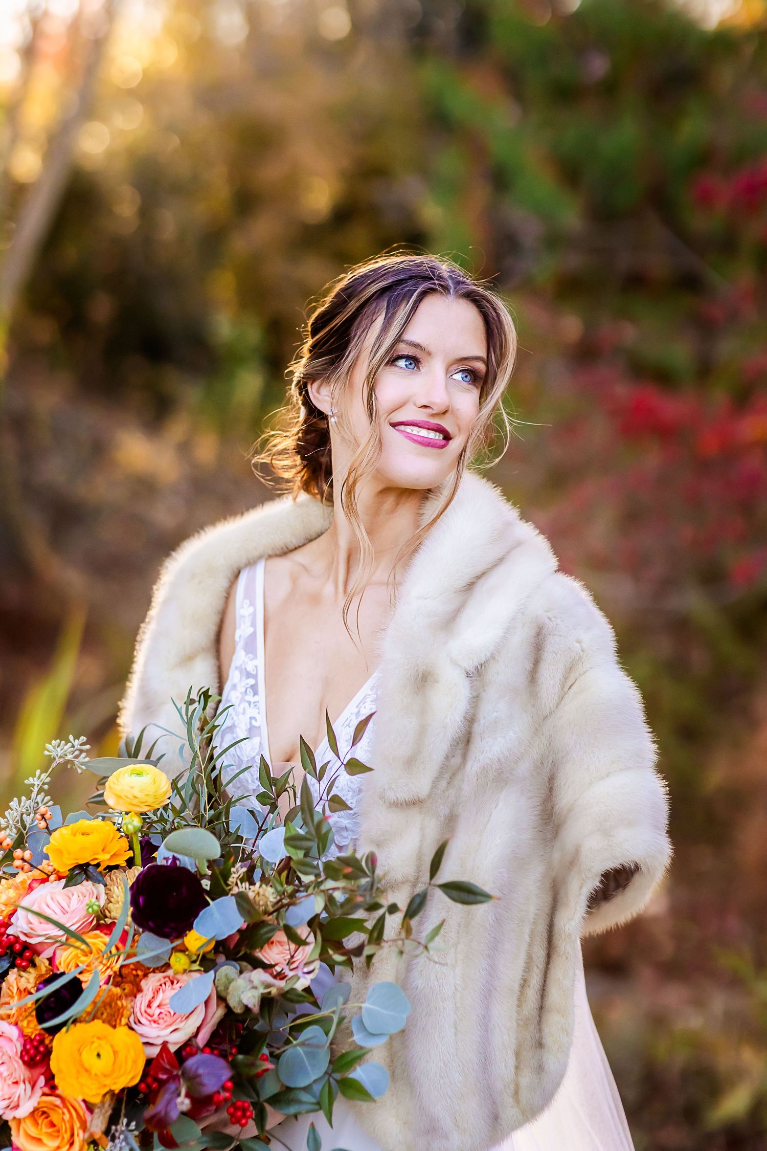 Bride in white fur wrap holds vibrant autumn bouquet with yellow and orange flowers against fall foliage background.