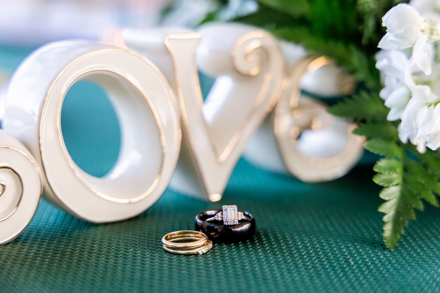 Wedding rings displayed with decorative white 'LOVE' letters and greenery on teal background.