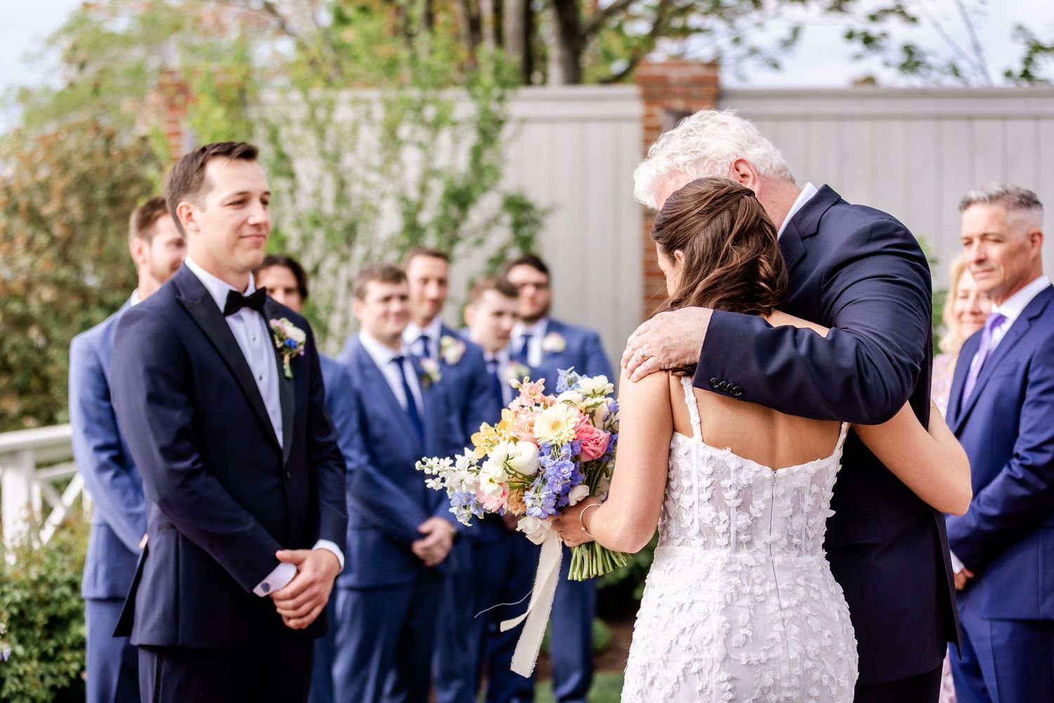 Wedding ceremony participants lined up outdoors with floral arrangements and white columns.
