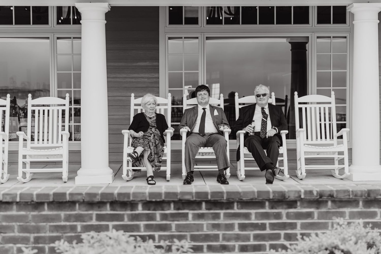 Wedding guests relaxing in white rocking chairs on a covered front porch.