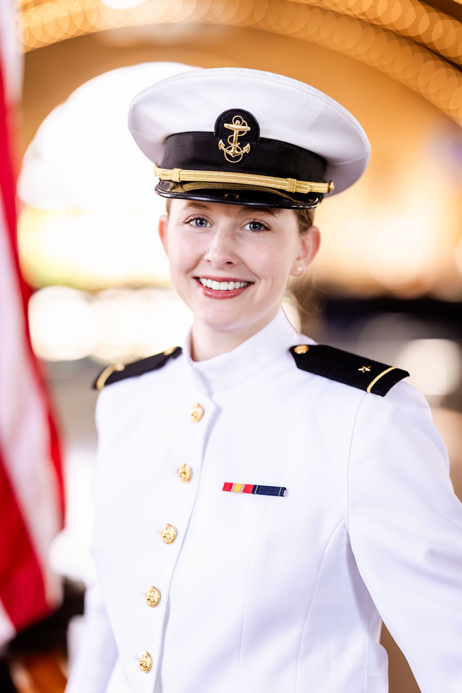 A naval officer in crisp white dress uniform with gold buttons and an officer's cap stands by an American flag.