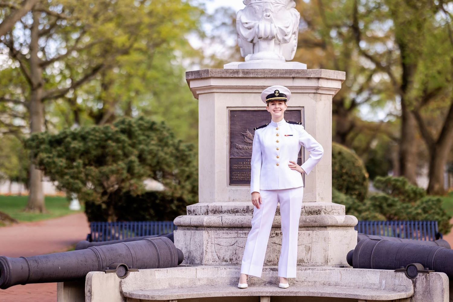 A person in white naval dress uniform stands at attention near a stone monument in a park setting.