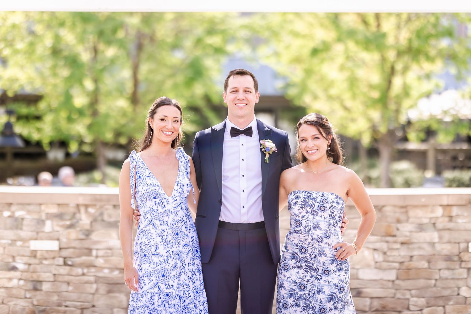 Wedding party members pose in blue floral dresses and formal attire against stone wall.
