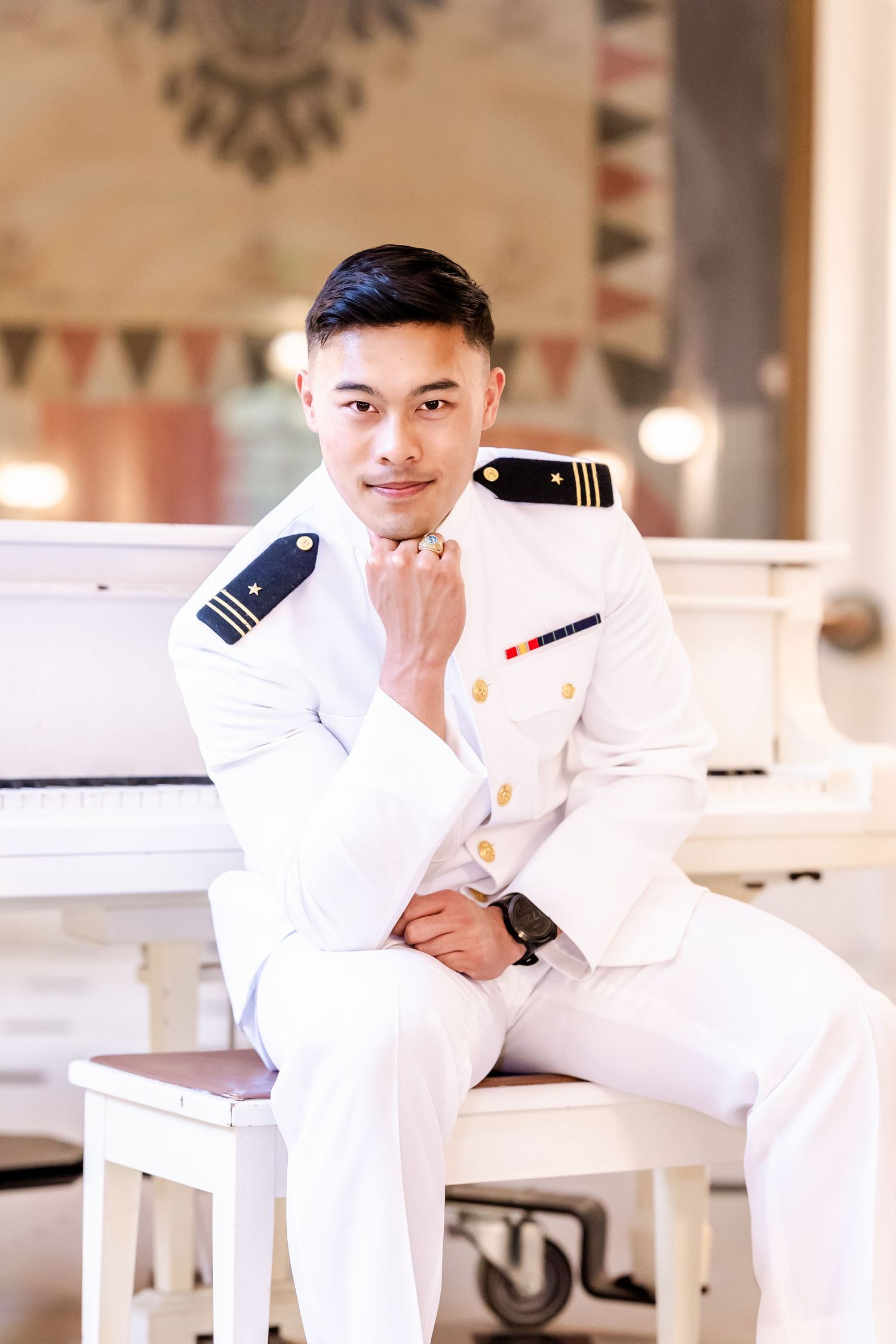 A naval officer in white dress uniform poses thoughtfully on a piano bench in an elegant interior setting.