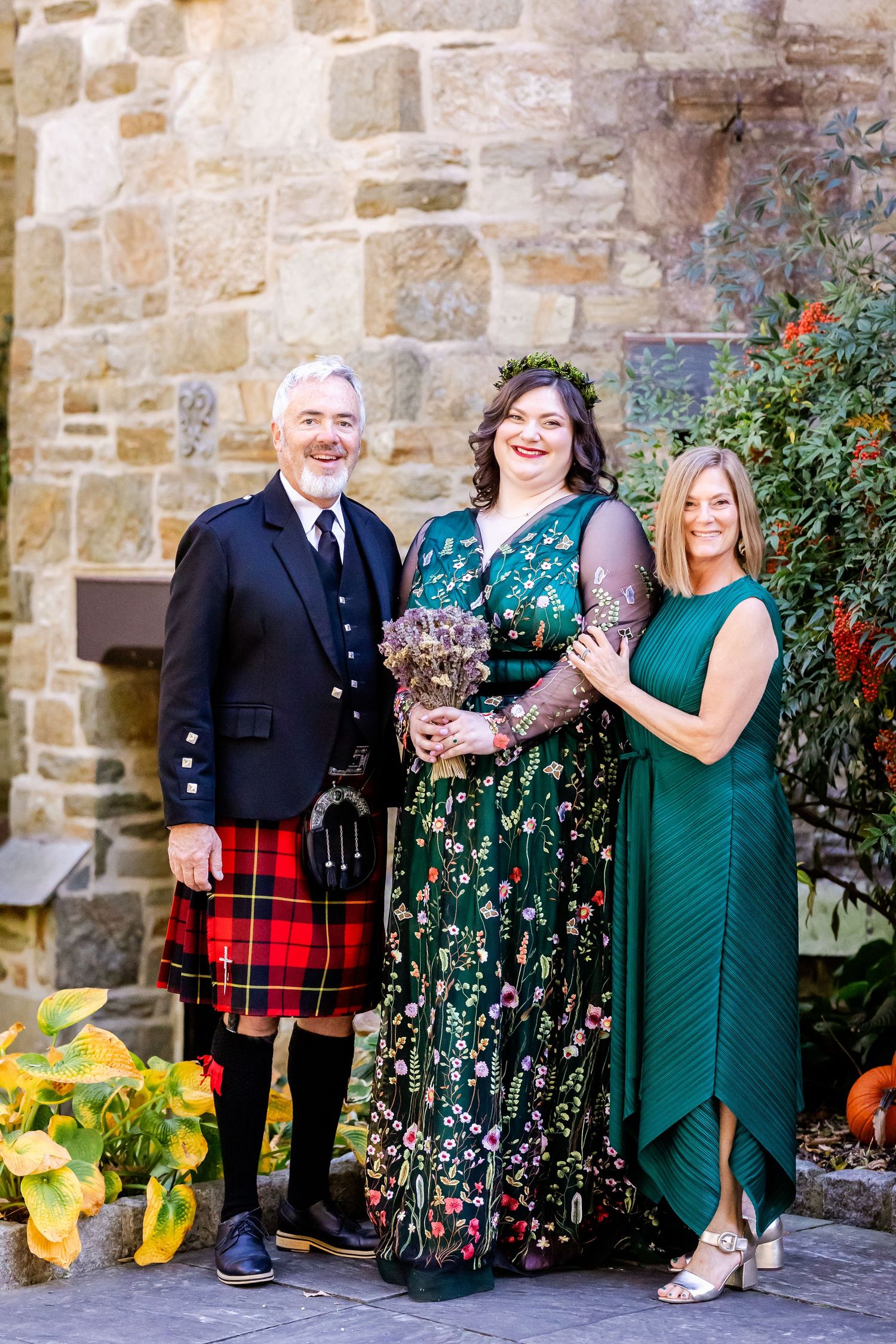 Wedding party members pose against stone wall, featuring traditional Scottish kilt and formal attire.