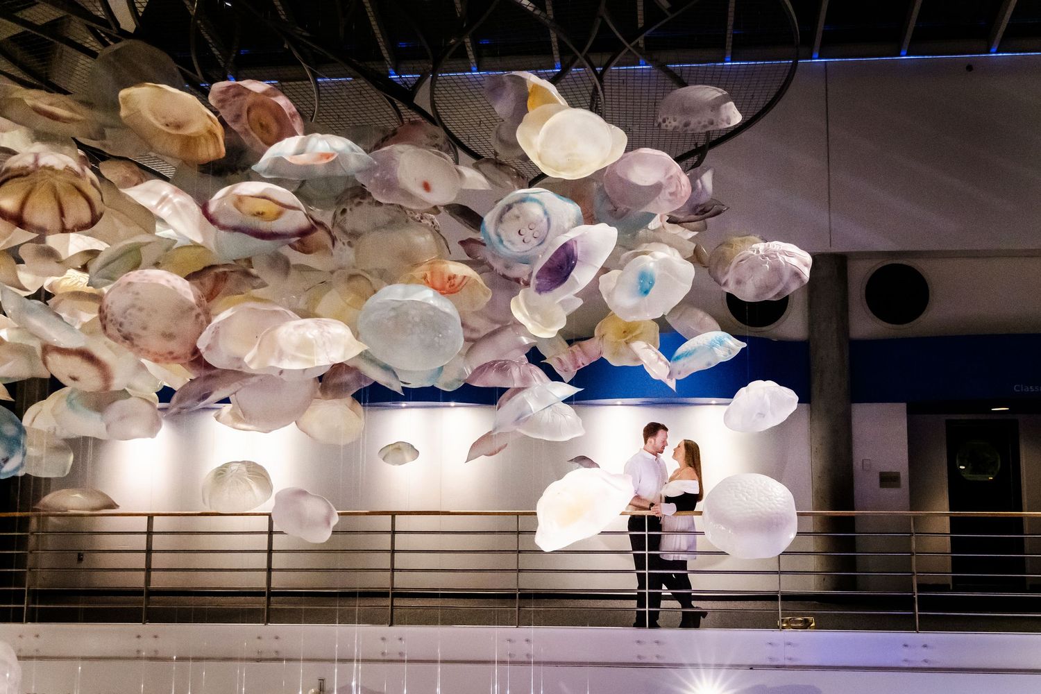 A couple embraces beneath an artistic ceiling installation of white hanging shells.