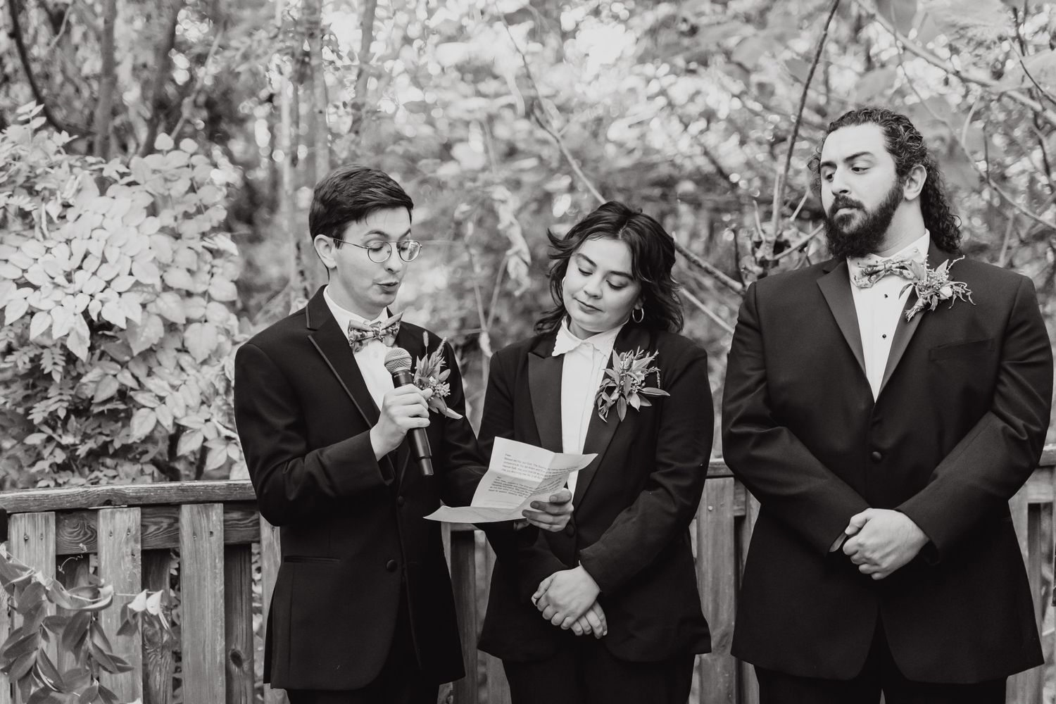 Black and white photo of a wedding ceremony taking place on a wooden deck in a garden setting.