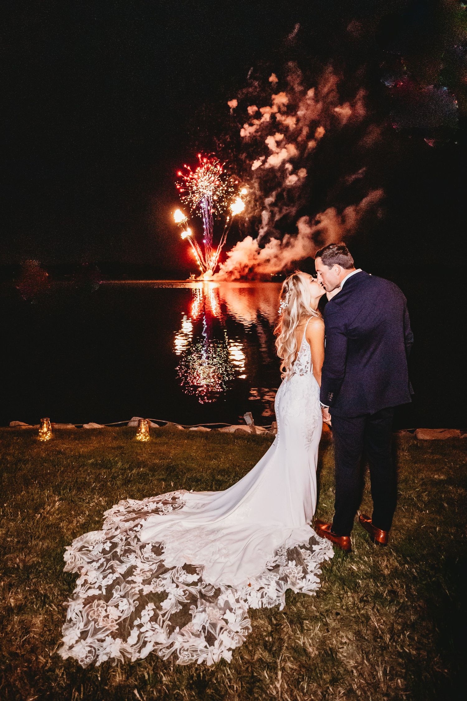Newlyweds share romantic moment by lakeside as fireworks reflect on water during nighttime wedding celebration.