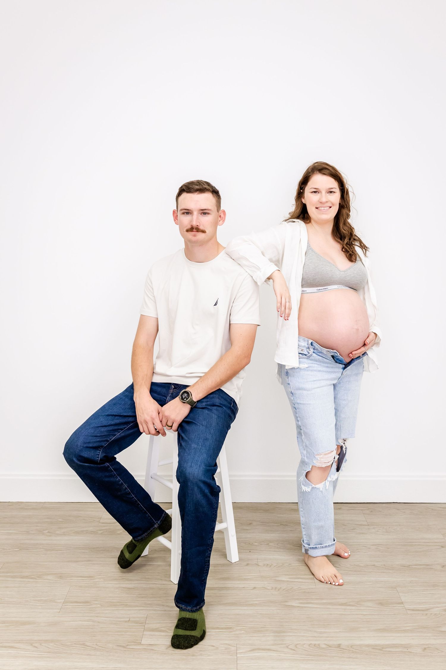 Casual studio maternity photo with couple in white and denim against minimalist background.