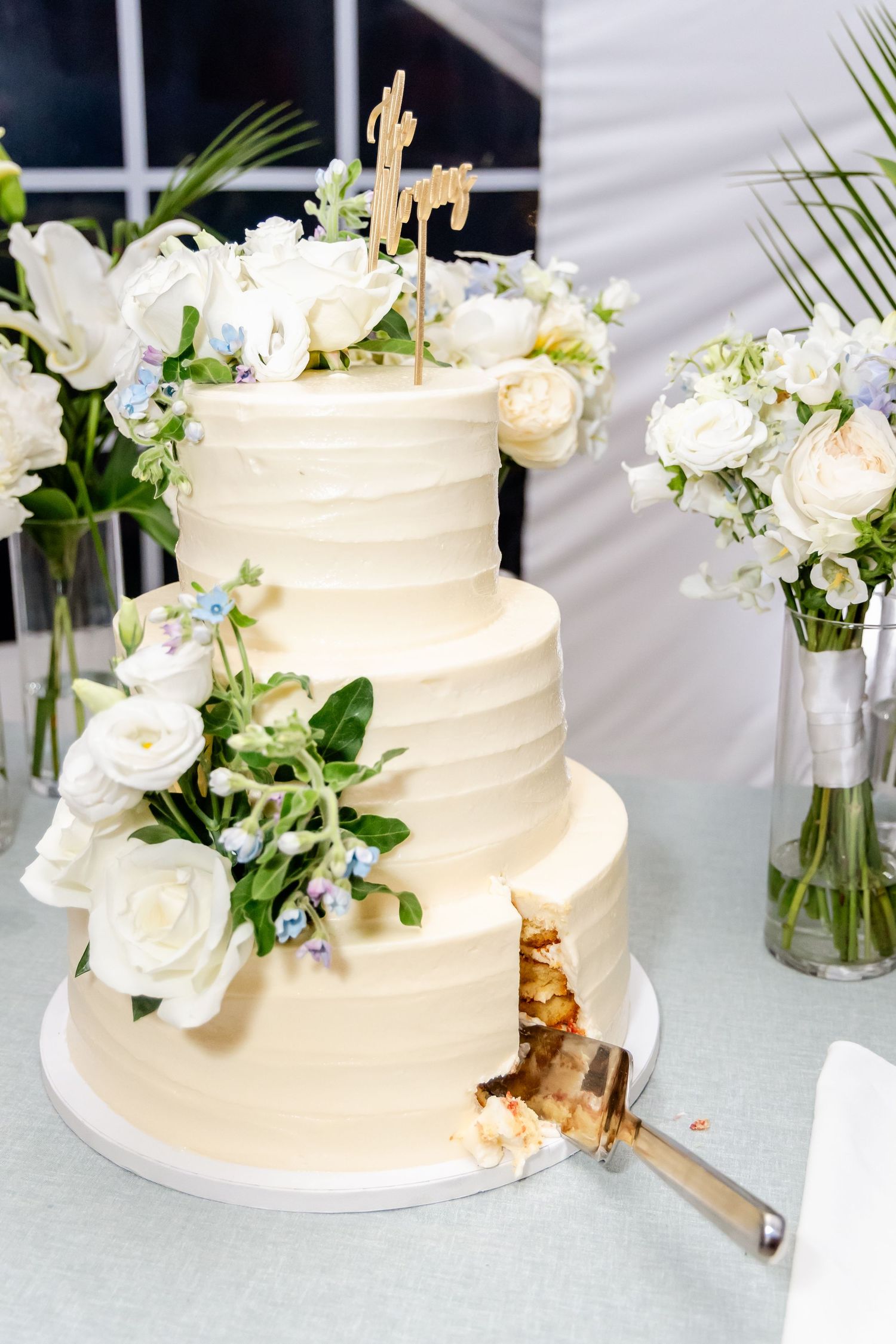 Three elegant white wedding cakes decorated with fresh flowers and greenery displayed on a light surface.