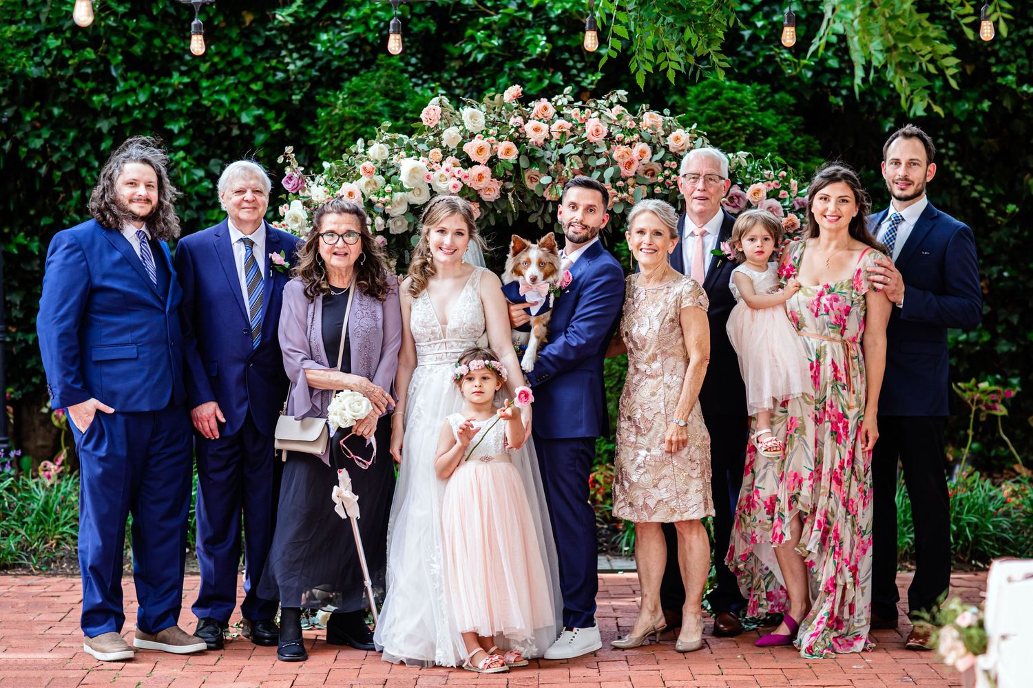 A group of formally dressed people stand together outdoors for a festive celebration with string lights and flowers in background.
