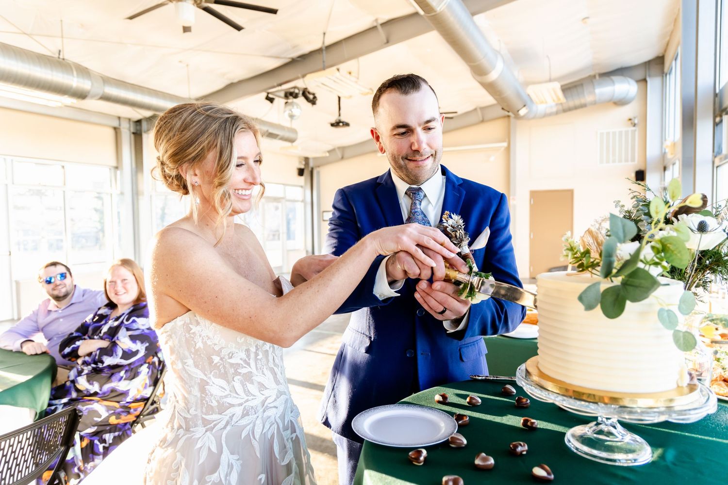 A couple in wedding attire cut their elegant white wedding cake together during their indoor reception celebration.