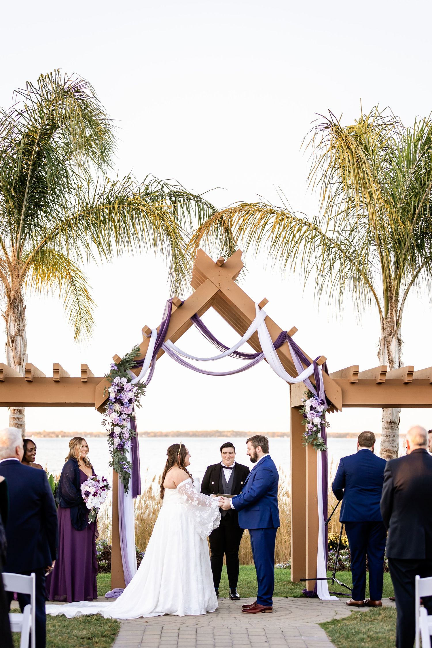 Outdoor wedding ceremony under decorative wooden arch with palm trees and water view.