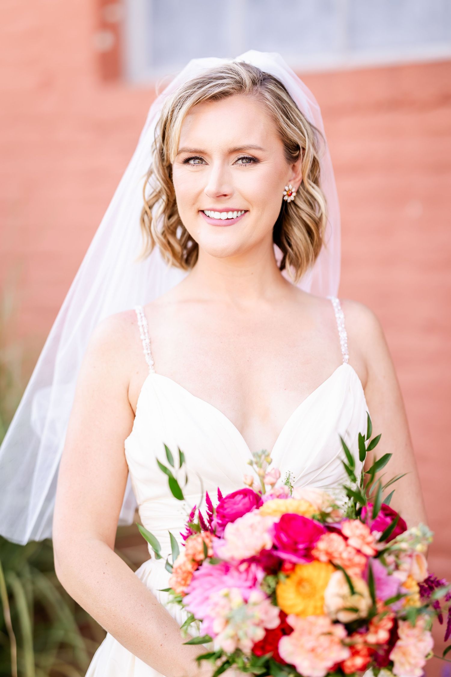 Bride in white dress holds vibrant pink and orange bouquet against brick wall backdrop.