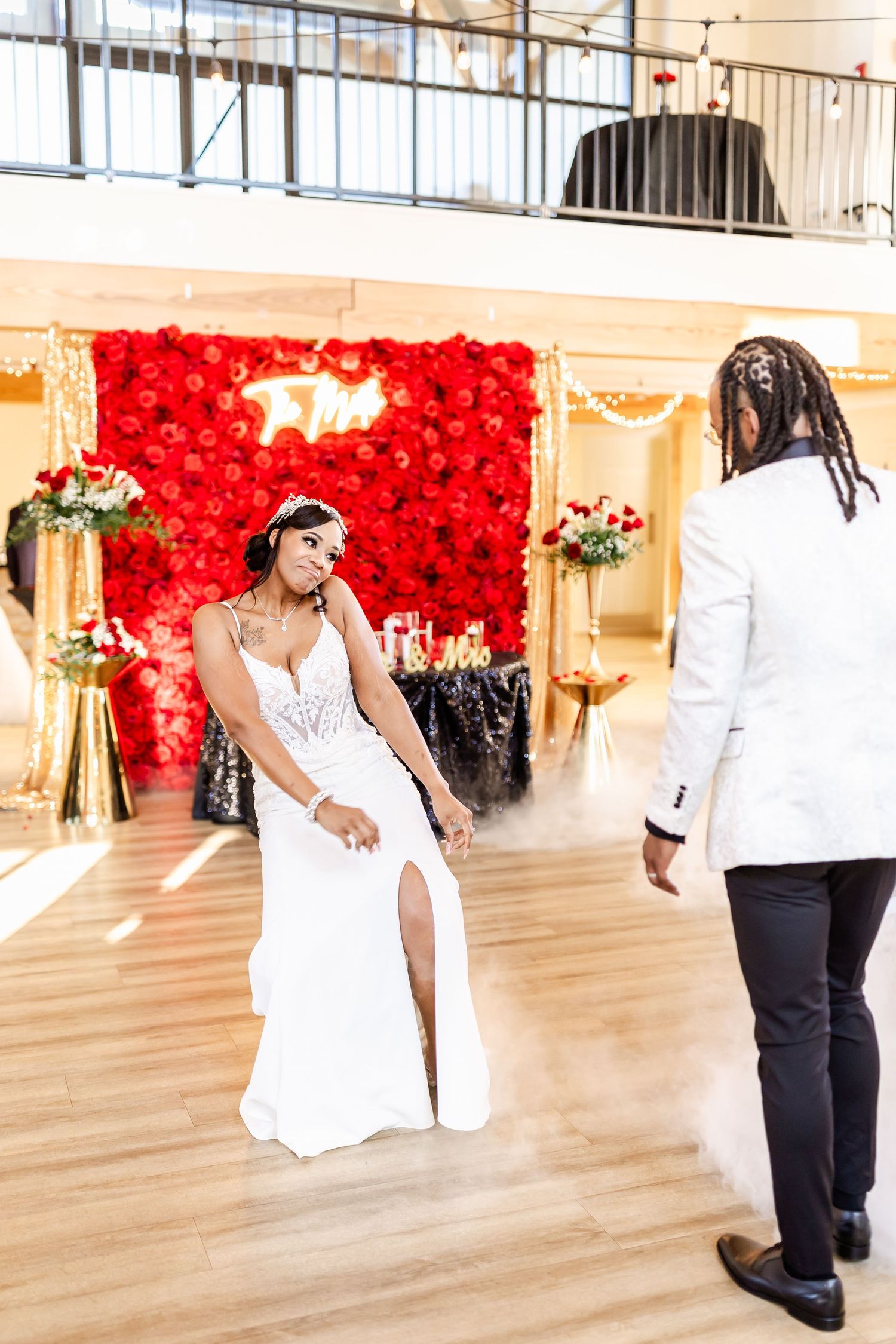 Wedding couple in white attire dance against a vibrant red rose backdrop at an elegant indoor venue.
