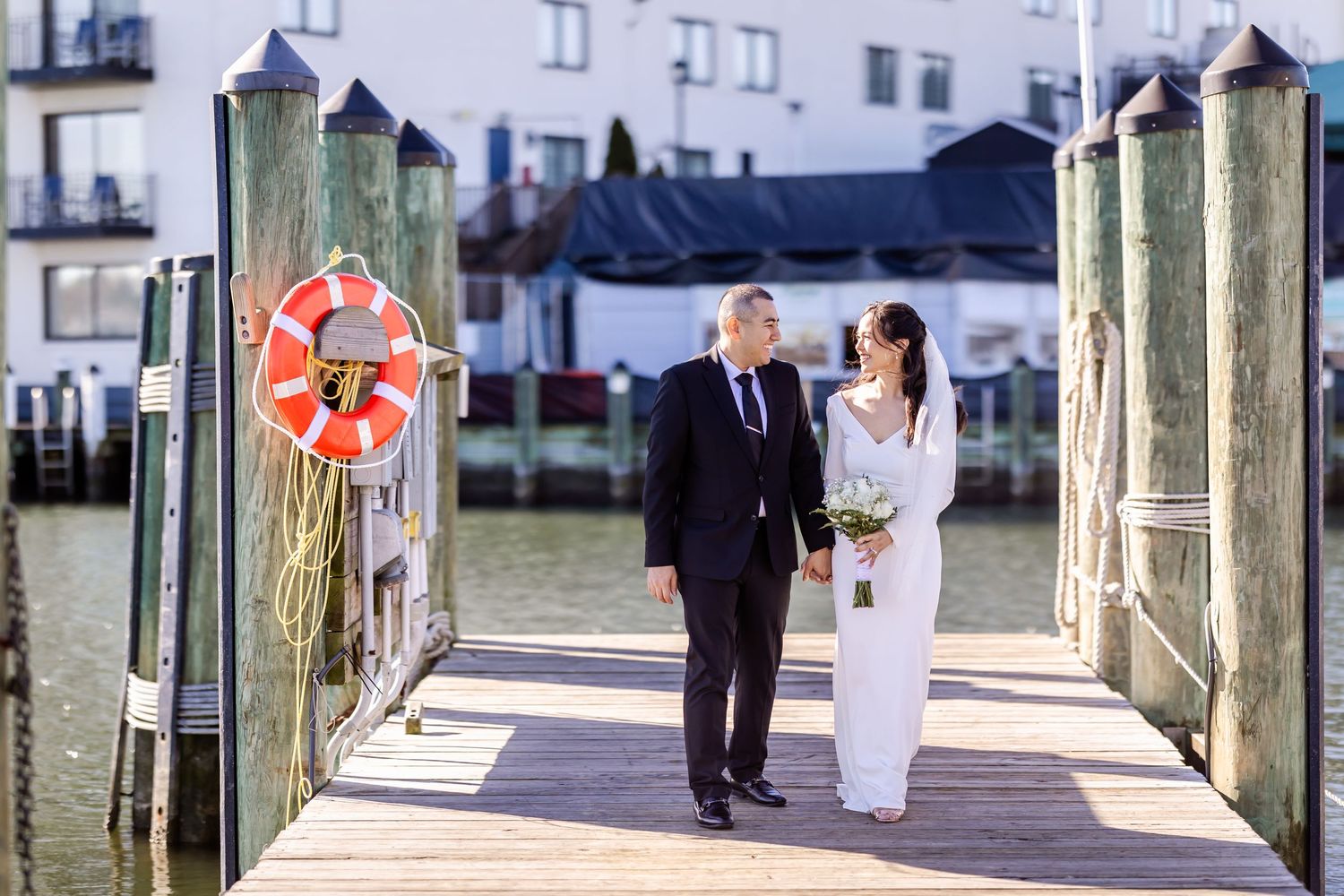 A couple walks along a waterfront dock with a life preserver and buildings in the background.