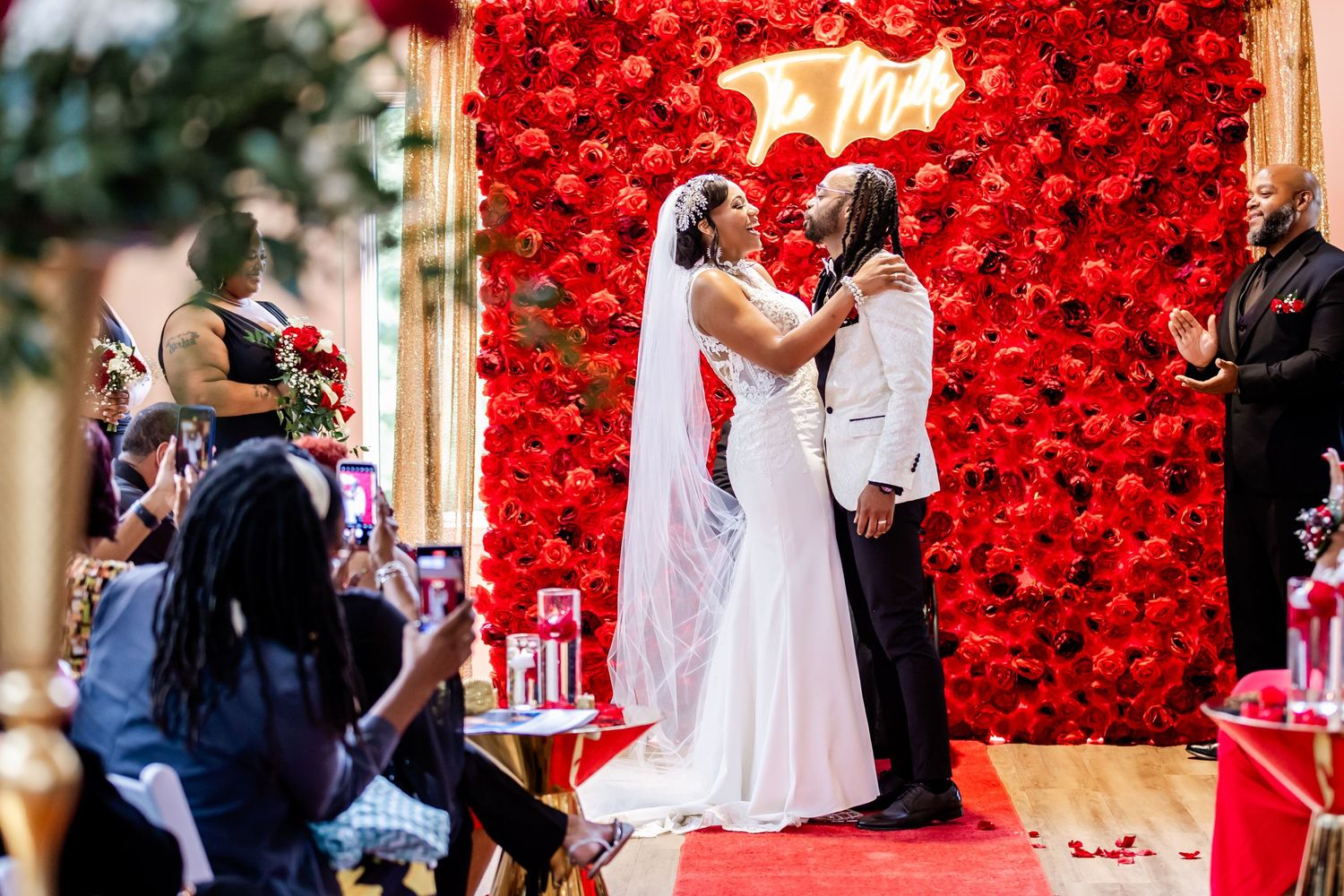 Romantic ceremony moment in front of dramatic red flower wall with onlookers.