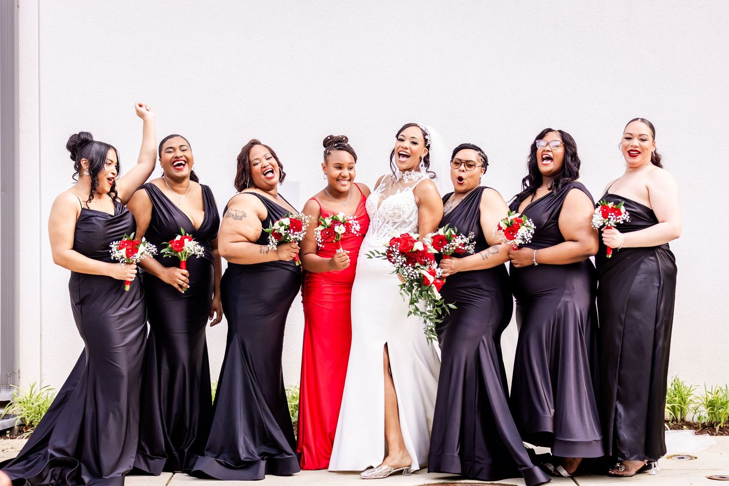 Wedding party poses in elegant black dresses with one in red, holding bouquets against white wall.