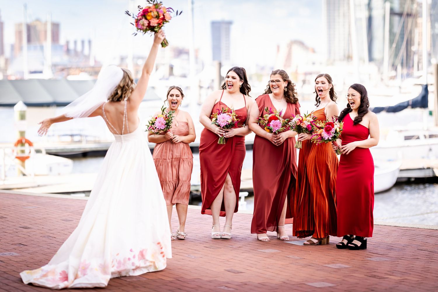 Wedding party in shades of red and coral poses on Baltimore harbor waterfront.