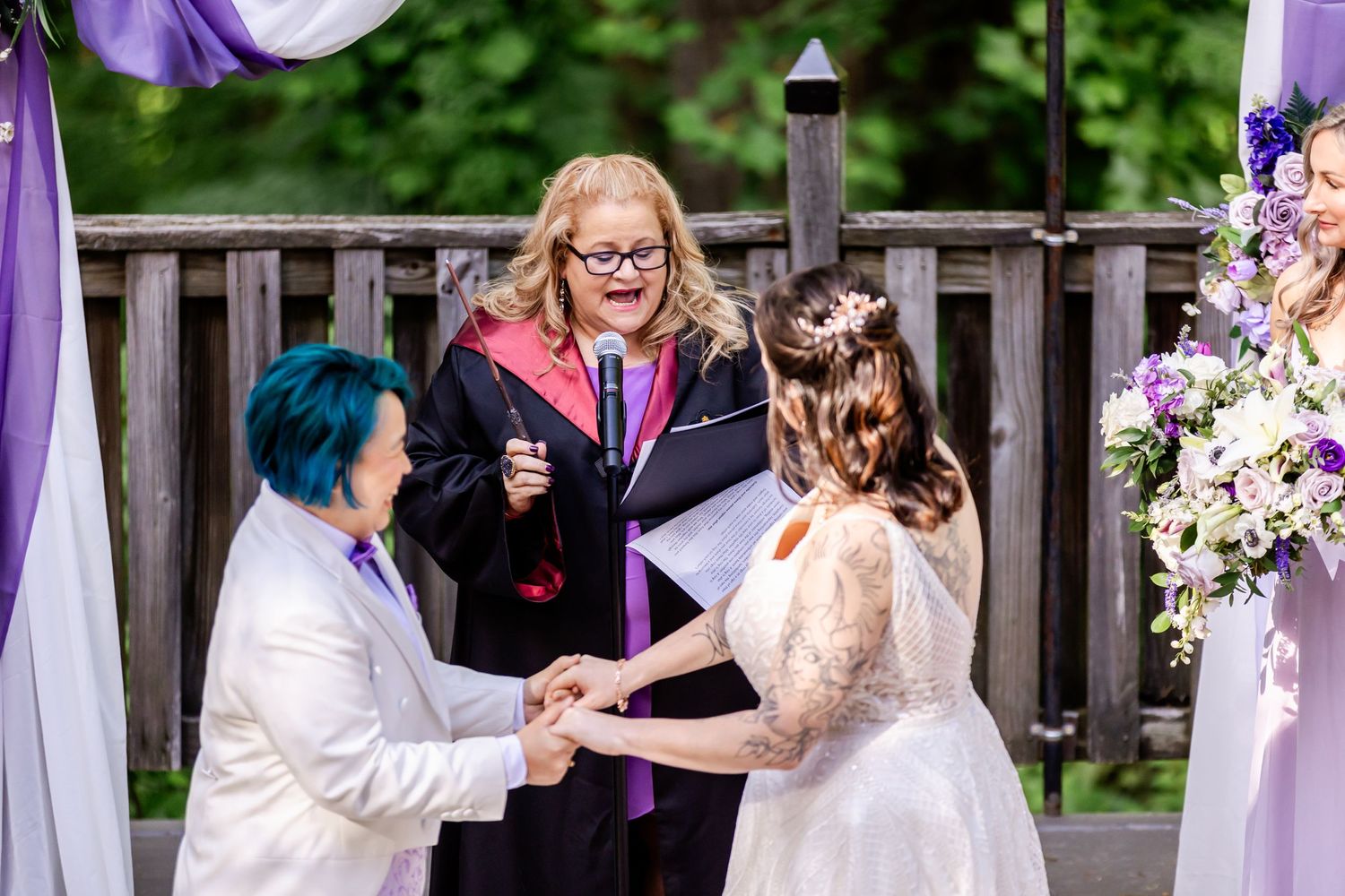 A wedding ceremony takes place on a wooden deck with purple and white floral decorations.