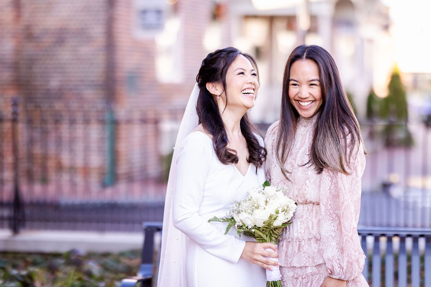 Two women in white and pink dresses share a joyful moment with a floral bouquet.