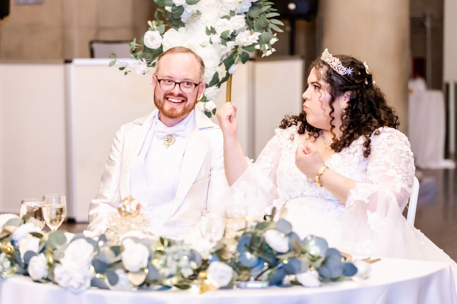 Couple shares laughter at head table during wedding reception with floral decorations.