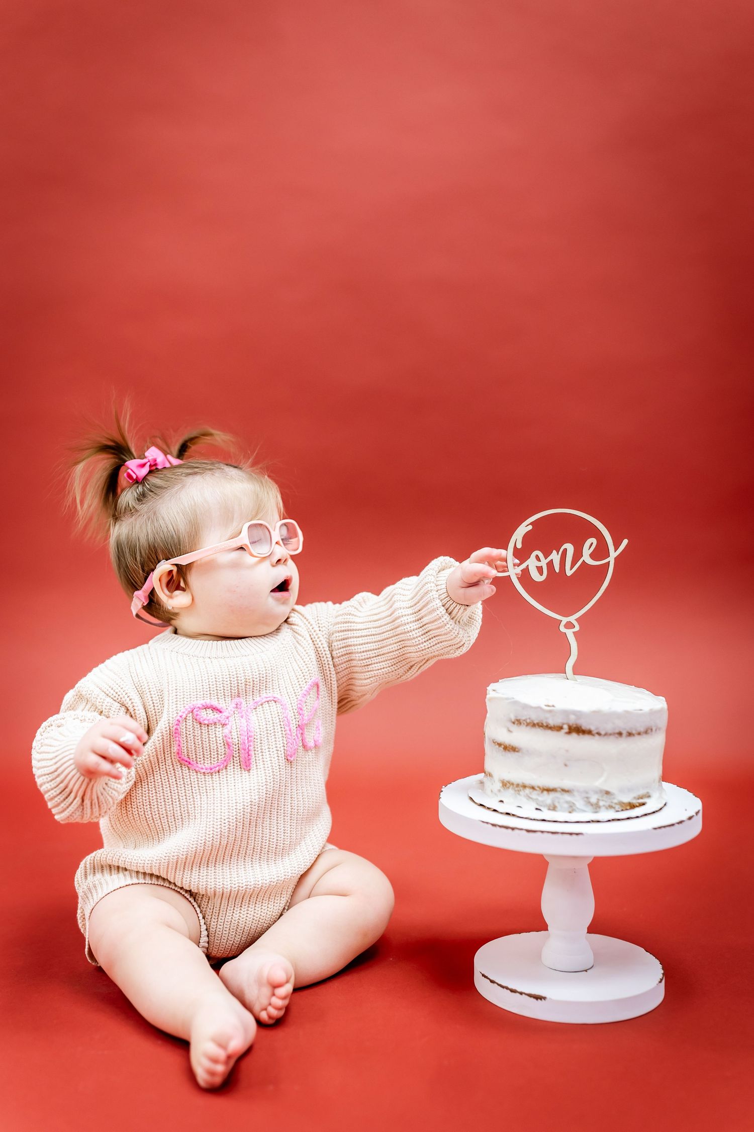 Baby in knit outfit reaches toward small white cake with 'one' topper on red backdrop.