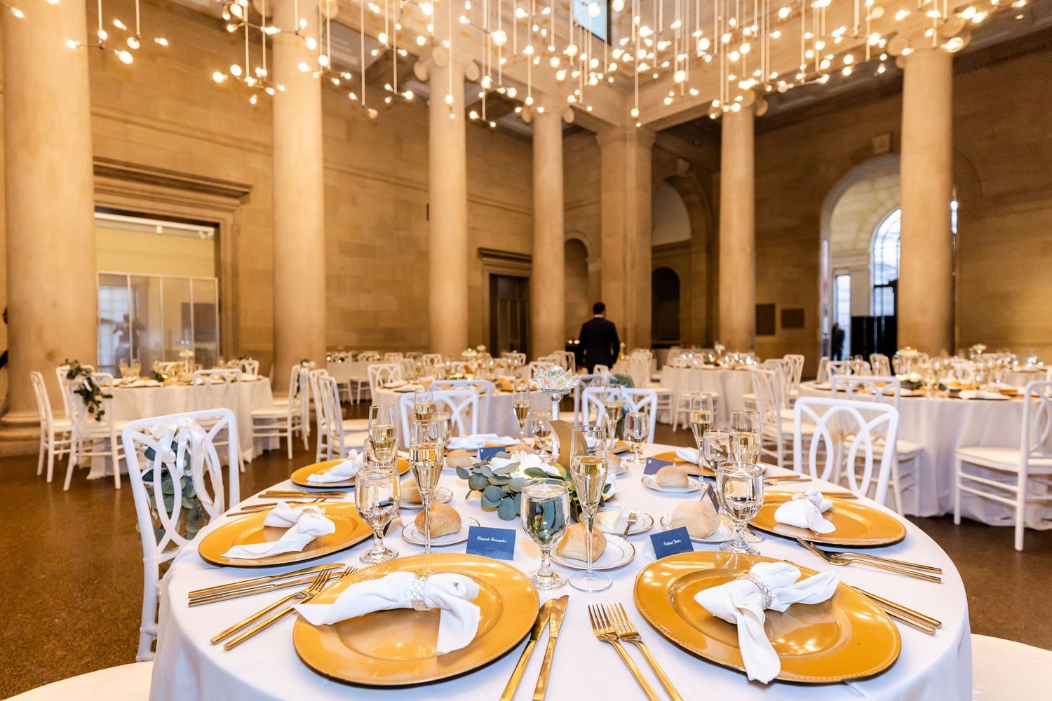 Grand ballroom wedding reception with hanging lights and gold charger plates on round tables.