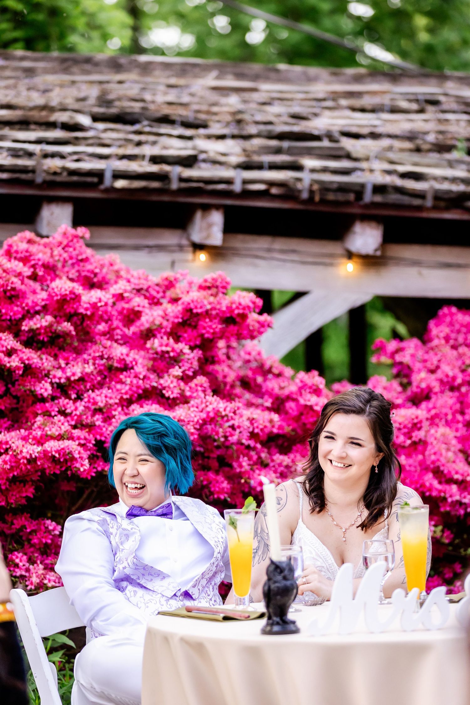 Two guests laugh together at a decorated outdoor table with bright pink azalea bushes in the background.