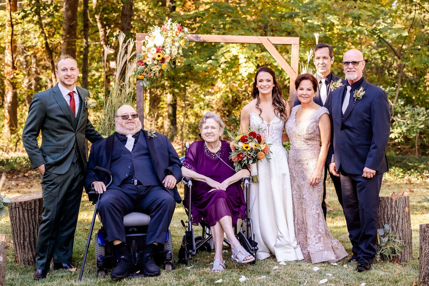 A formal wedding party portrait taken outdoors with members wearing purple, burgundy and navy attire.