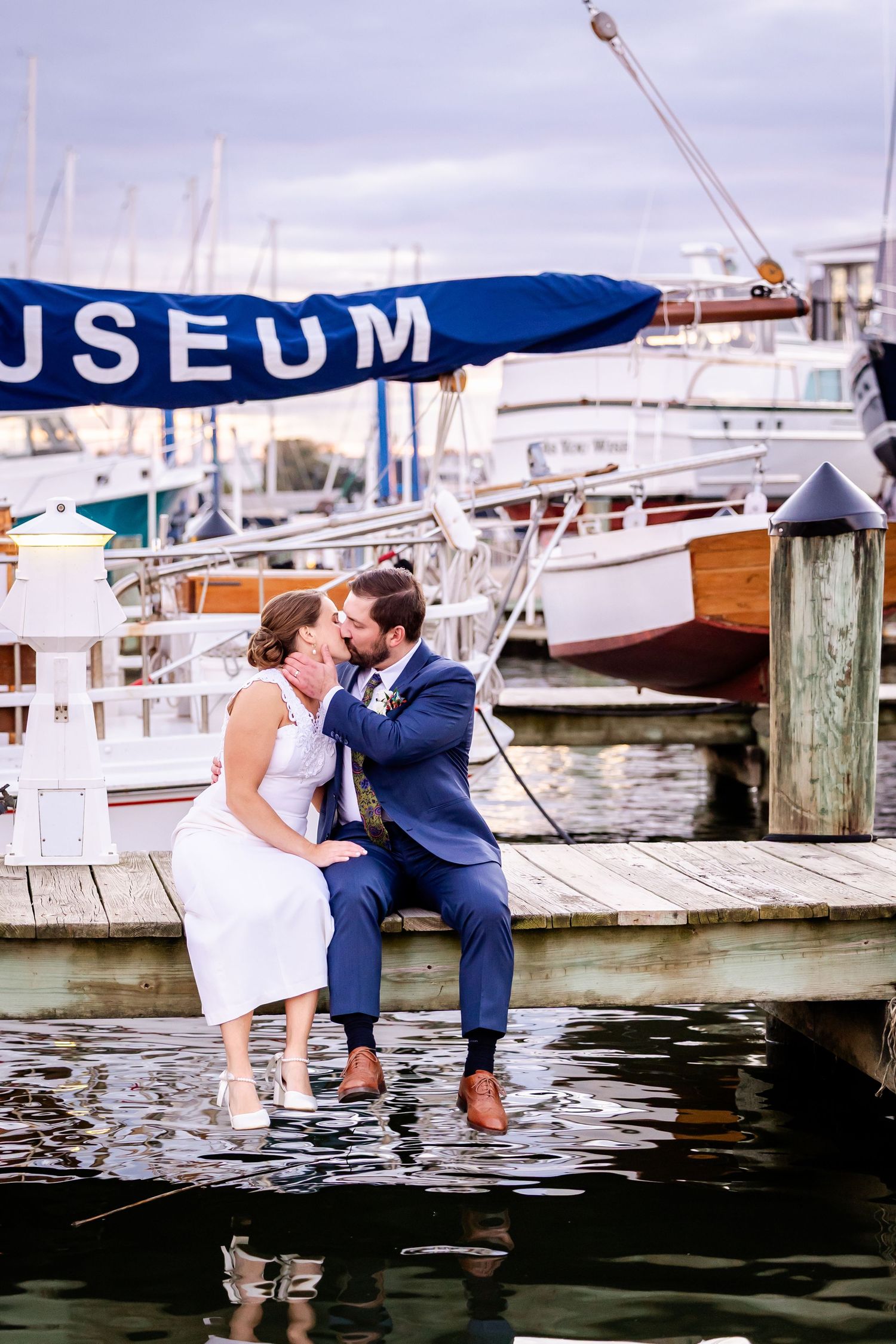 A romantic couple in formal attire share a kiss while sitting on a dock at a marina with boats in the background.