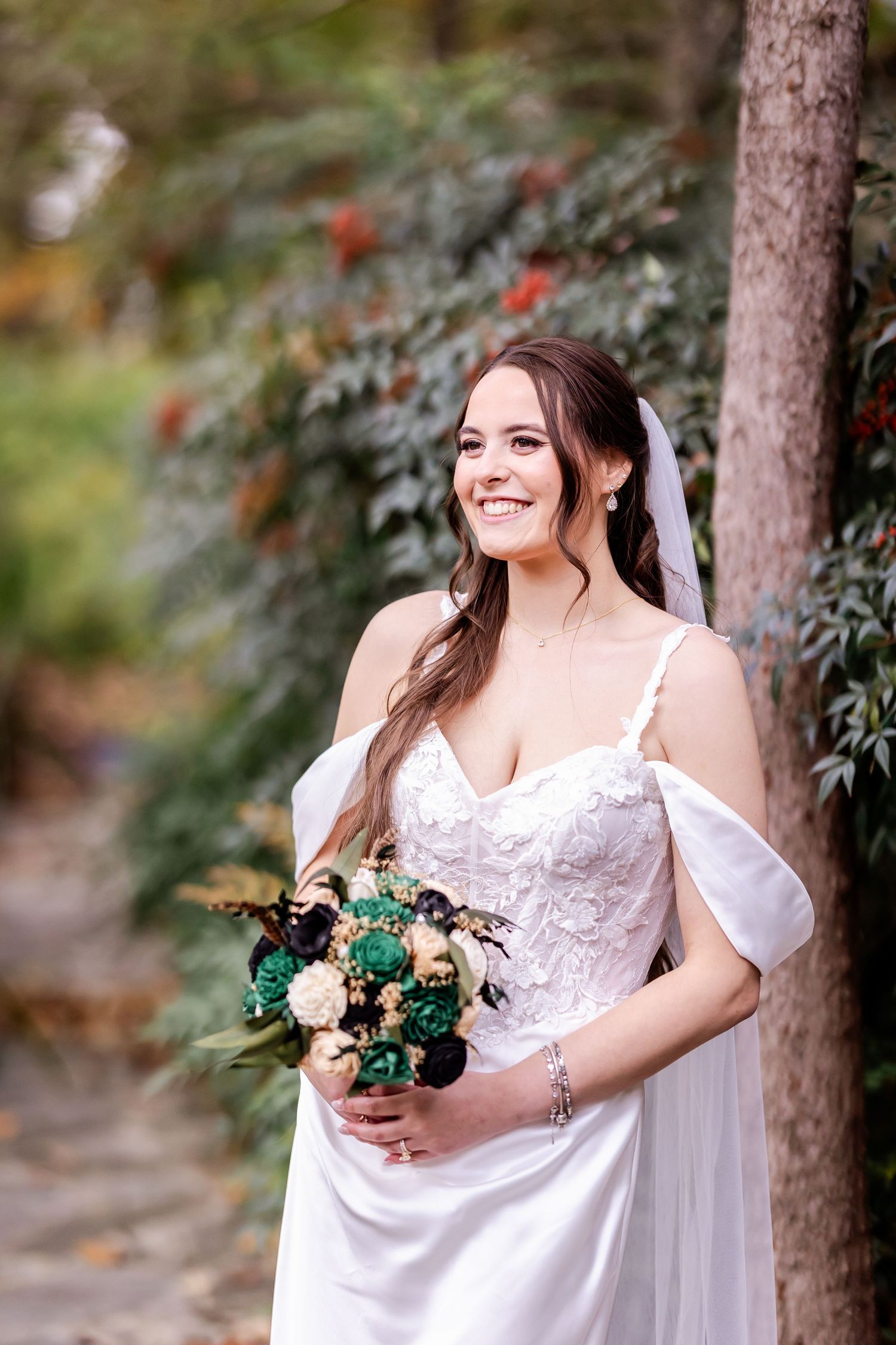A bride in a white lace wedding dress poses outdoors among trees while holding a romantic bouquet of flowers.