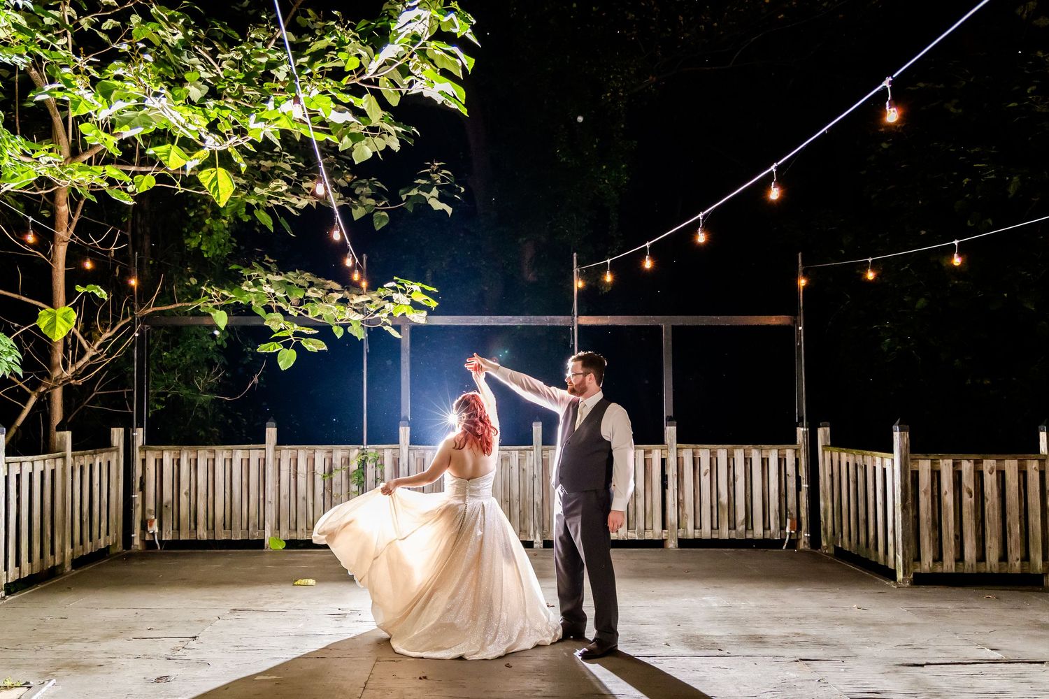 A couple shares a romantic dance moment under string lights on an outdoor wooden deck at night.
