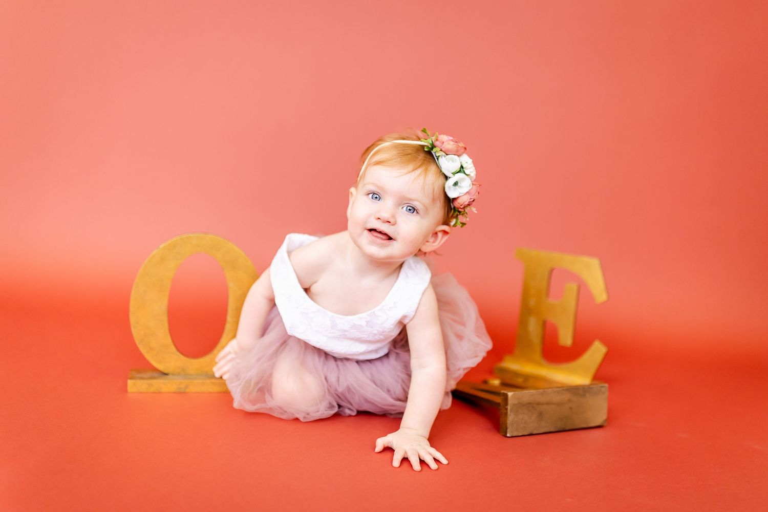 Baby in pink dress sits in white chair against coral background with playful expressions.