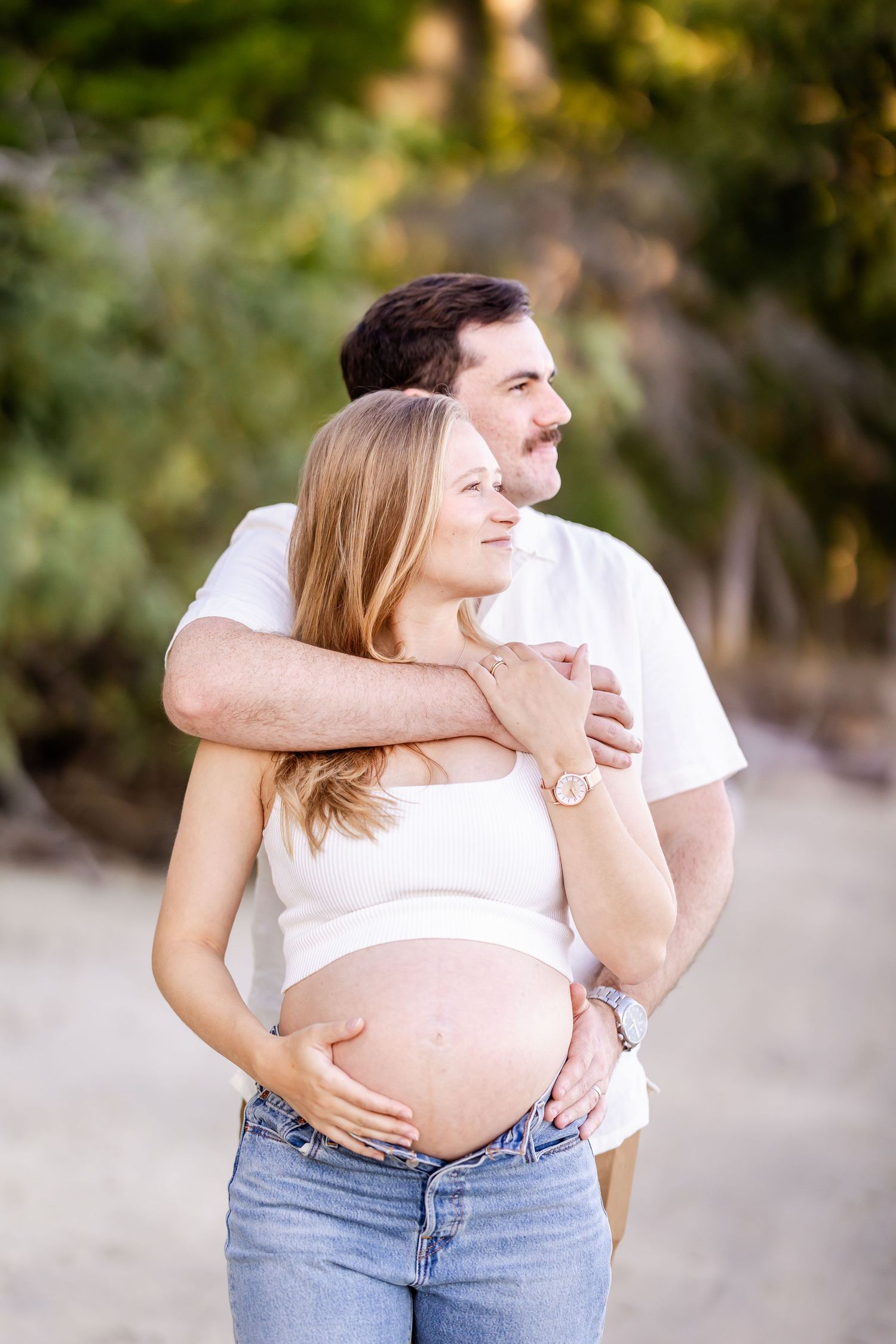 Expectant couple embracing outdoors in white shirts and jeans during sunset photoshoot.