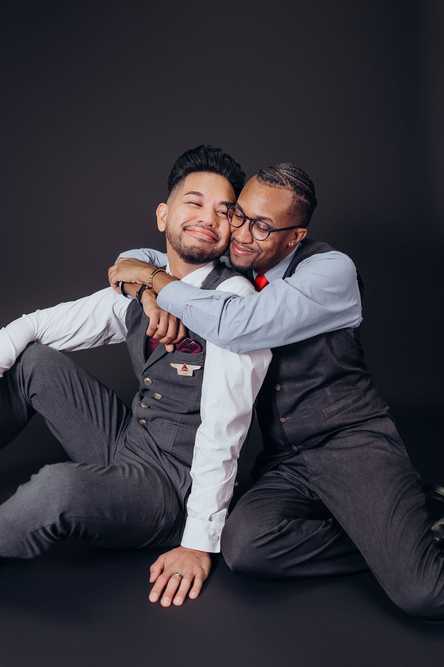 Two people in business attire embrace playfully while lying down against a dark background.