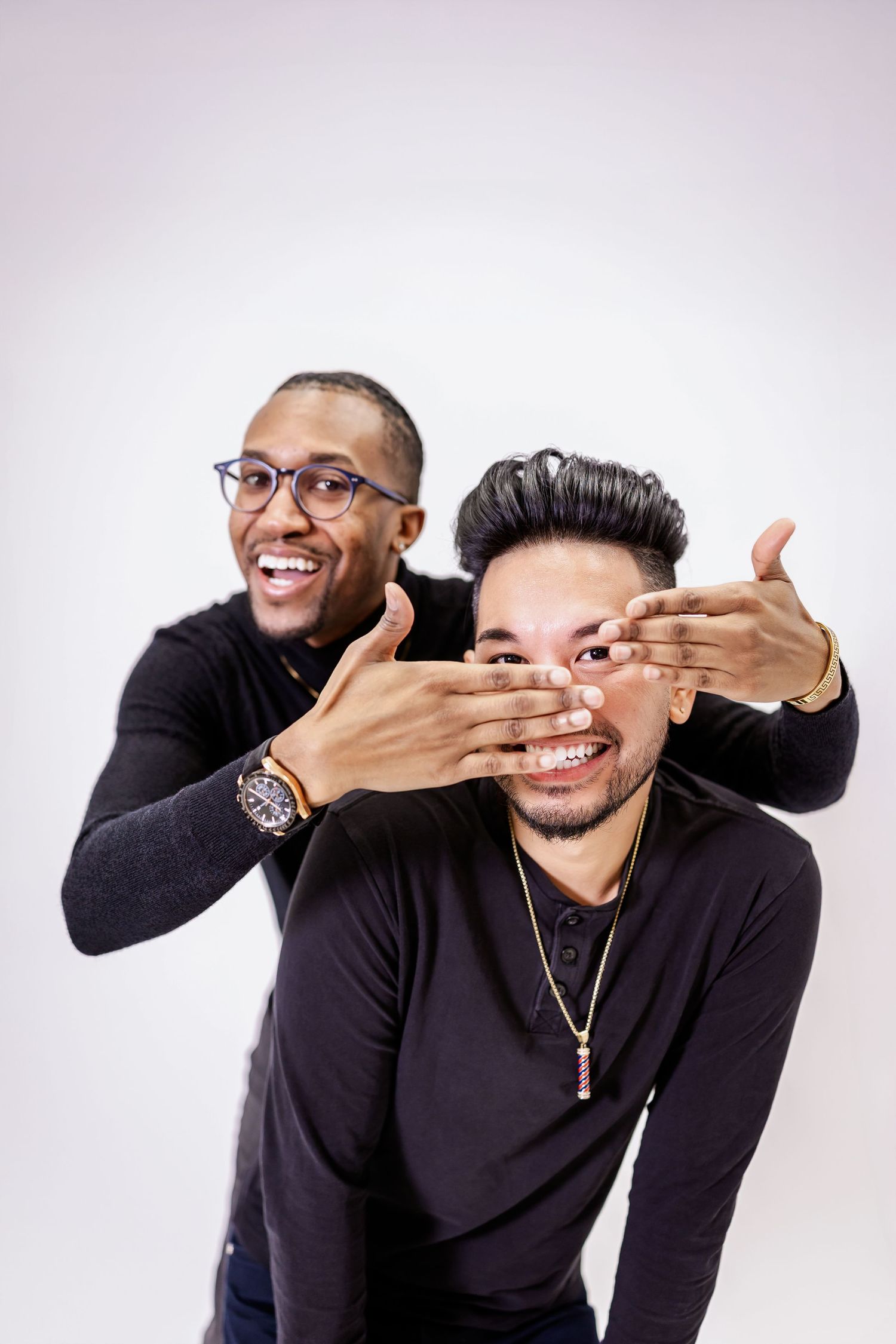 Two friends in black shirts playfully pose together against a light background, with one covering the other's eyes.
