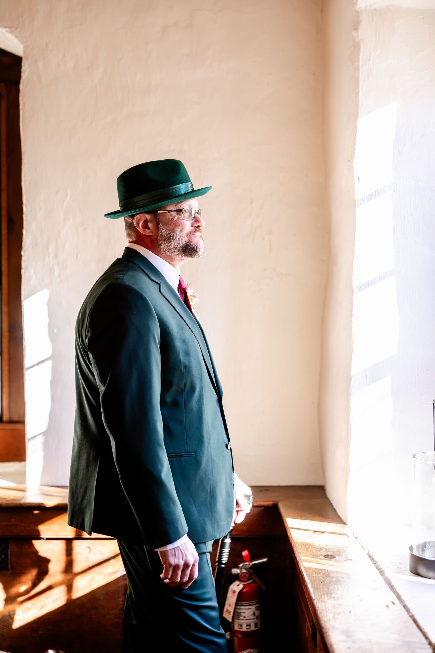 A person in a dark suit and green hat stands in profile against a beige wall in sunlight.