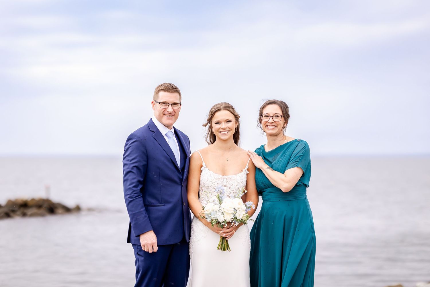 Wedding party members stand together on a cloudy beach in formal attire with the ocean visible in the background.