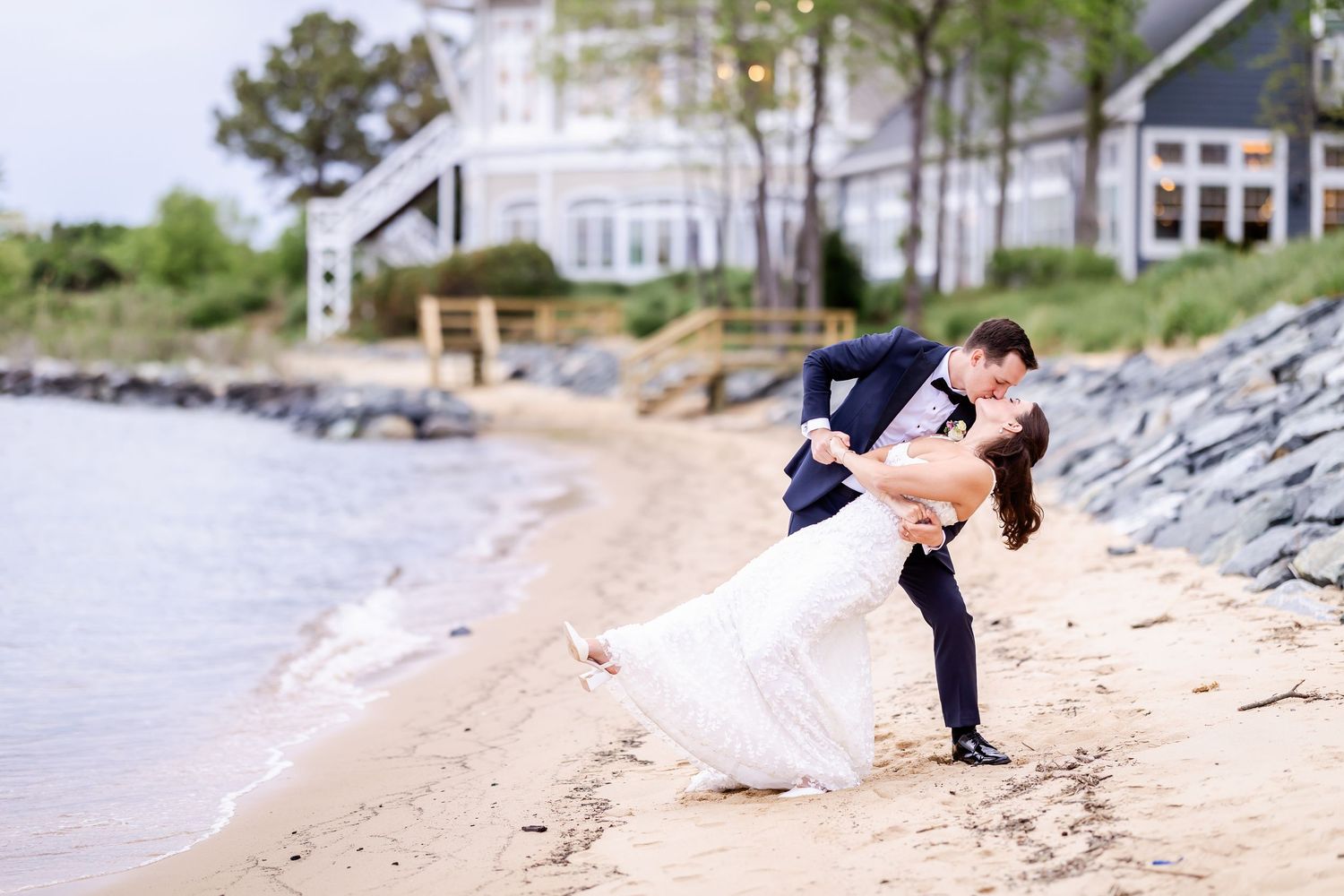 A romantic dip kiss on the beach with coastal homes in the background.
