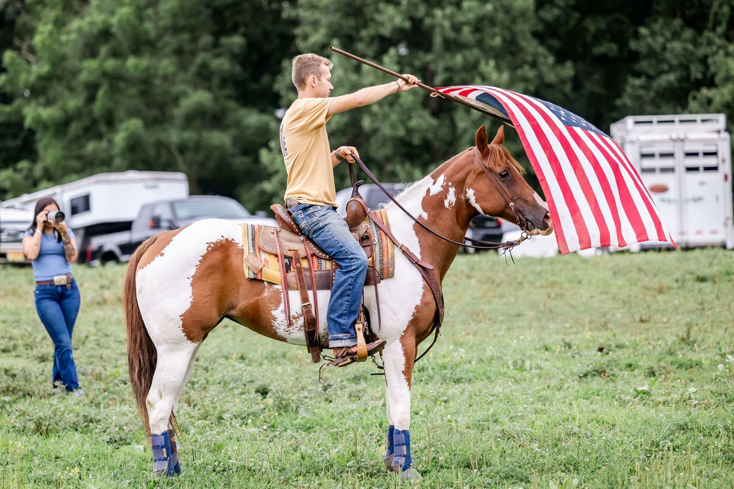 Equestrian rider carrying American flag while riding paint horse in grassy field.