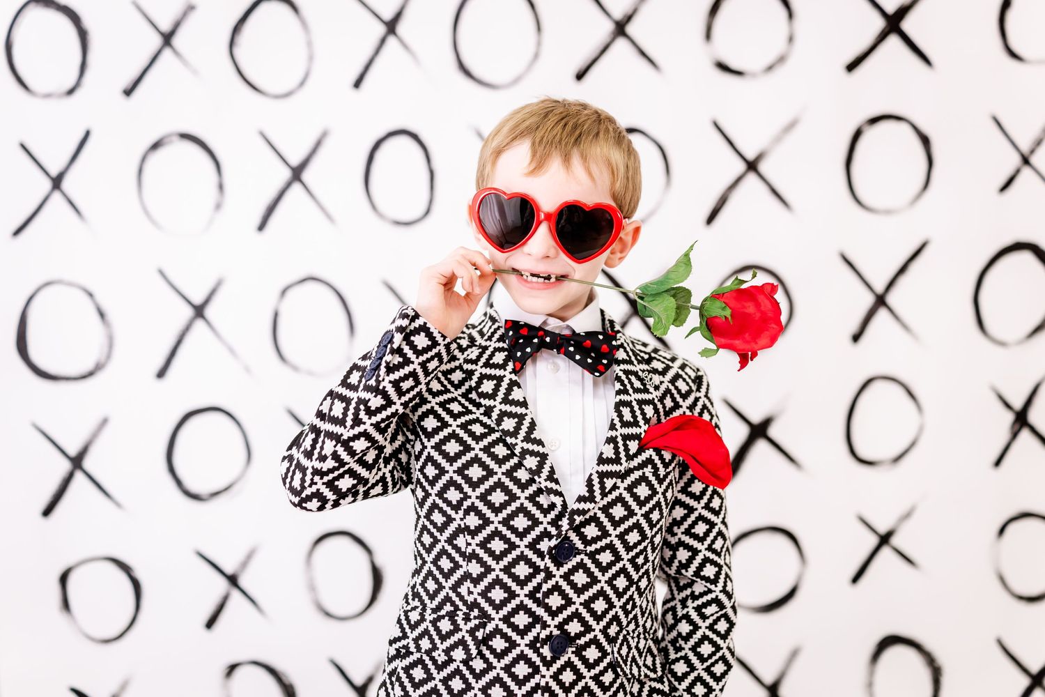 Small figure in red sunglasses and patterned jacket holds a red rose against black and white X's and O's background.