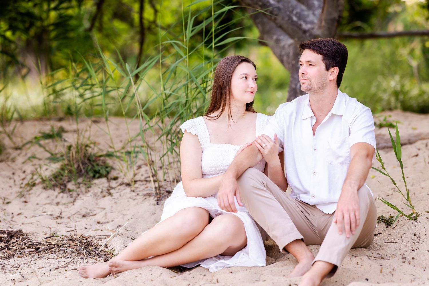 A couple wearing white clothing sits together on a sandy beach during a romantic outdoor photography session.