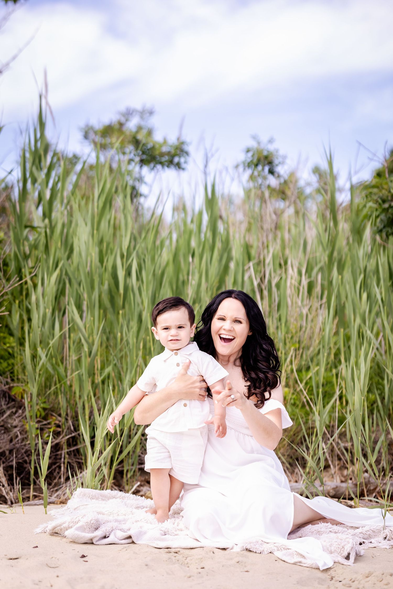 Two people in white outfits sit together and laugh joyfully on a sandy beach surrounded by tall grass on a sunny day.