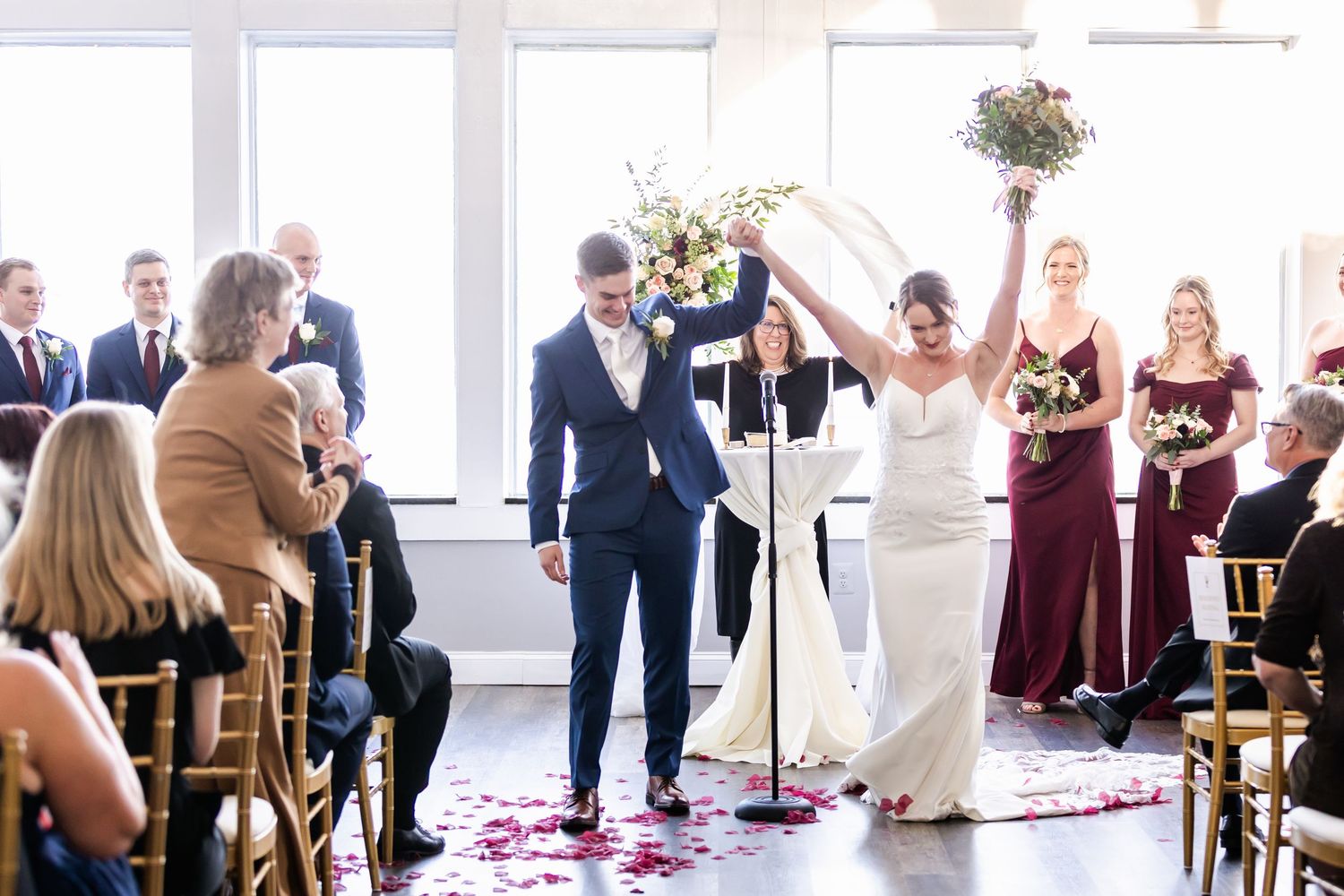 Wedding reception setup in bright room with guests seated at tables and rose petals on floor.