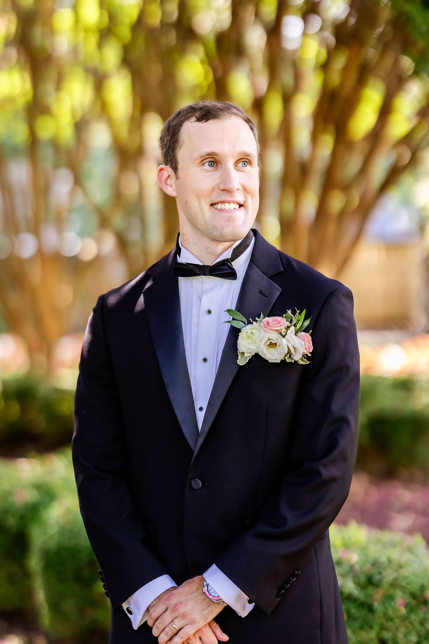 Elegant groom in classic black tuxedo and bowtie with boutonniere smiles in outdoor garden setting.