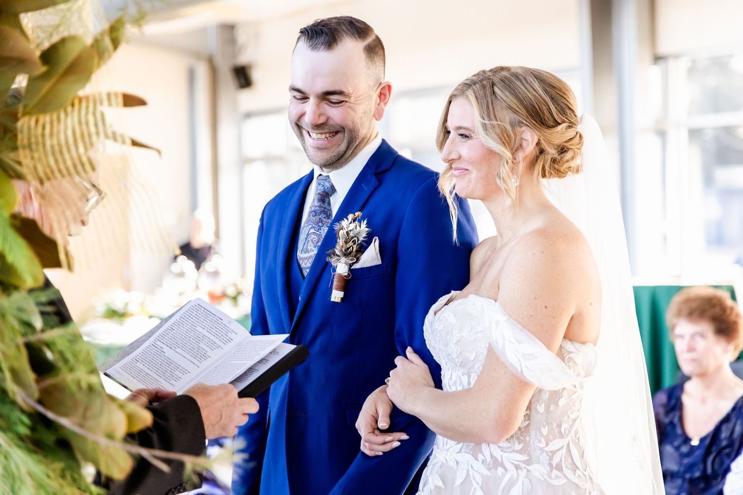A couple share a joyful moment during their wedding ceremony in a bright indoor venue with greenery.