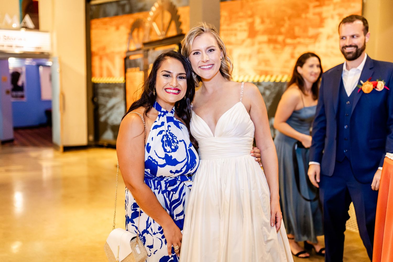 Two people pose together at a wedding reception with warm lighting and decorative walls behind them.