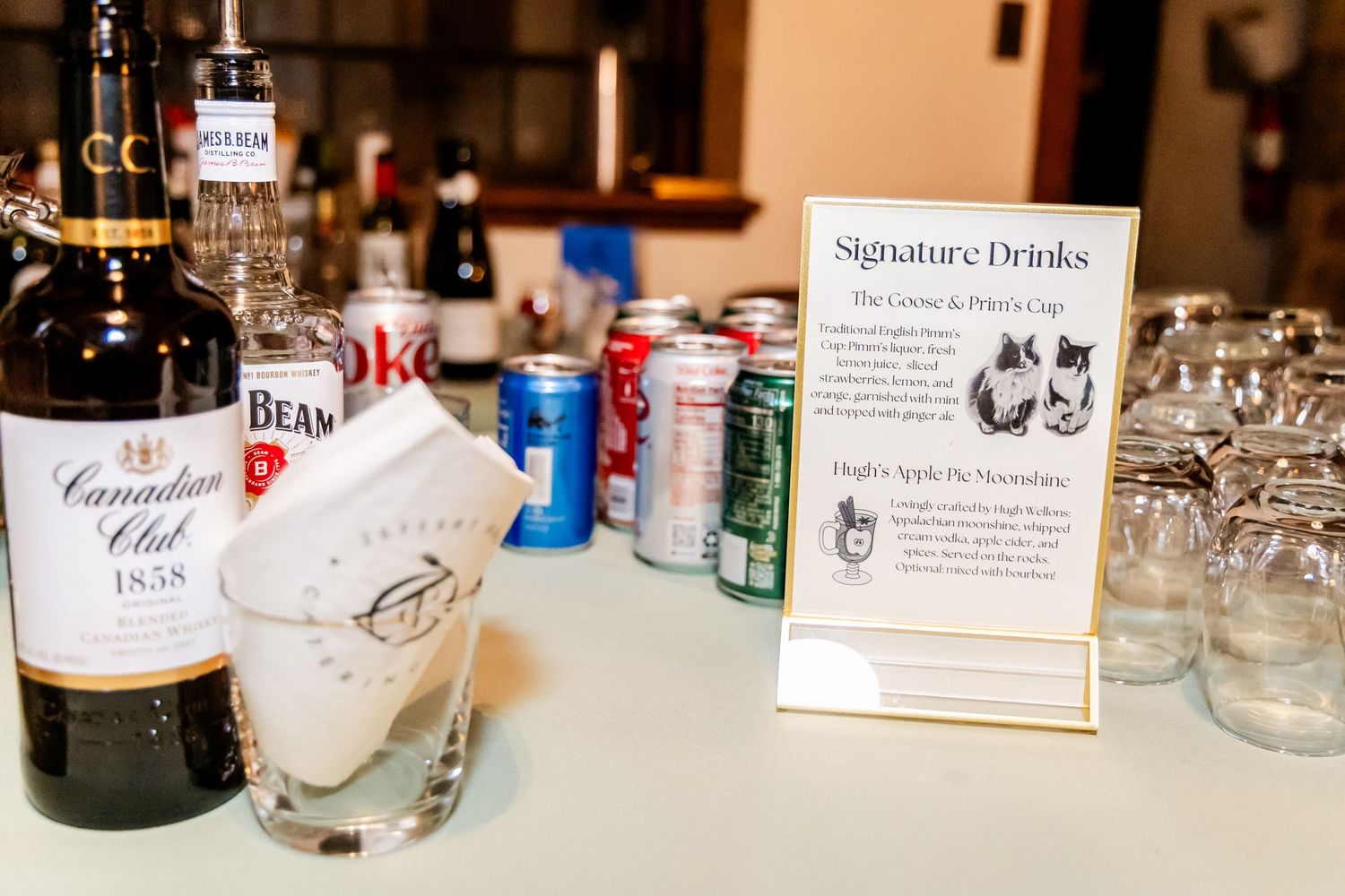 Wedding bar setup with signature drinks menu, liquor bottles and glassware on display.