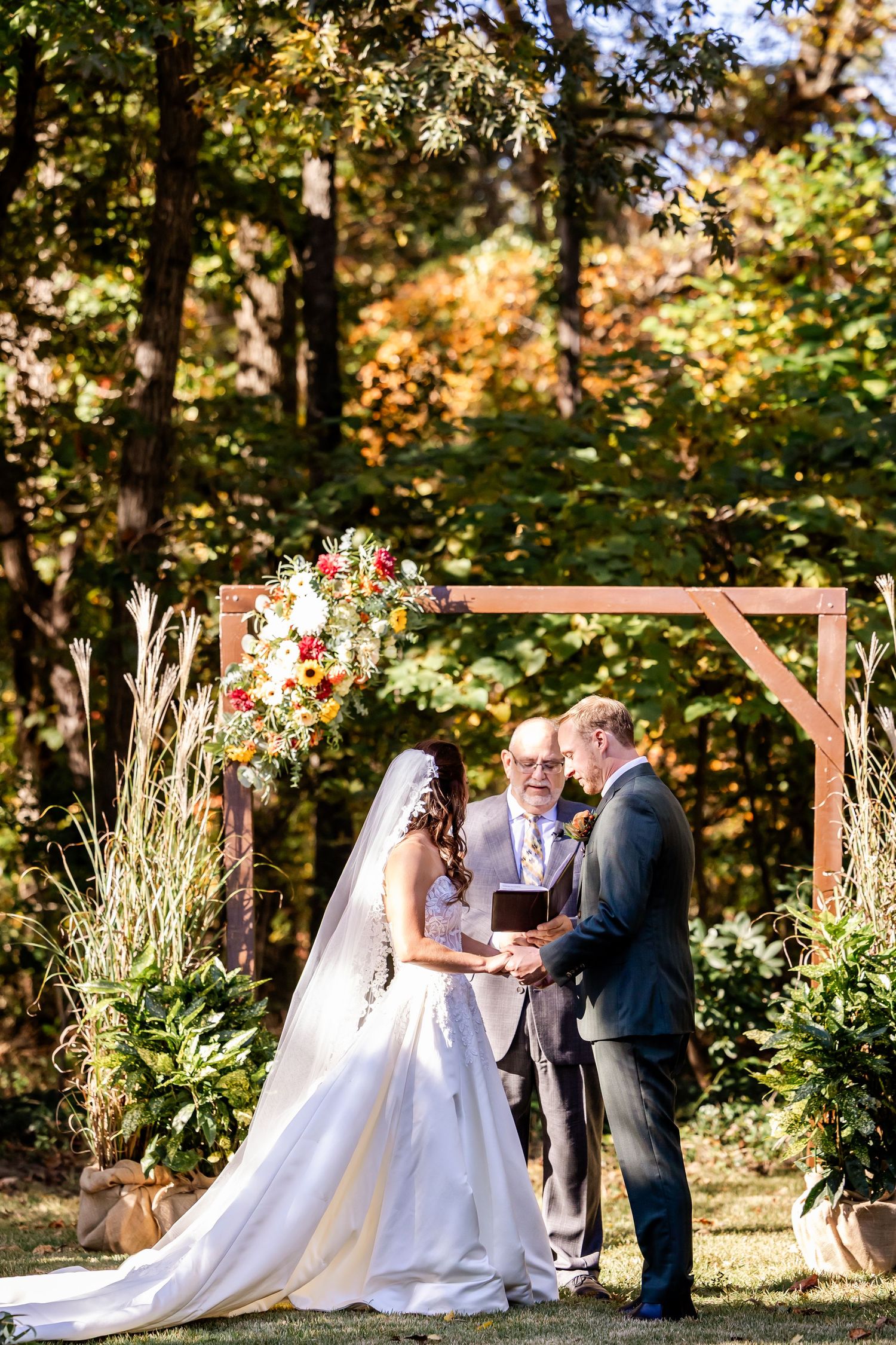 A romantic outdoor wedding ceremony takes place under a wooden arch surrounded by autumn foliage.
